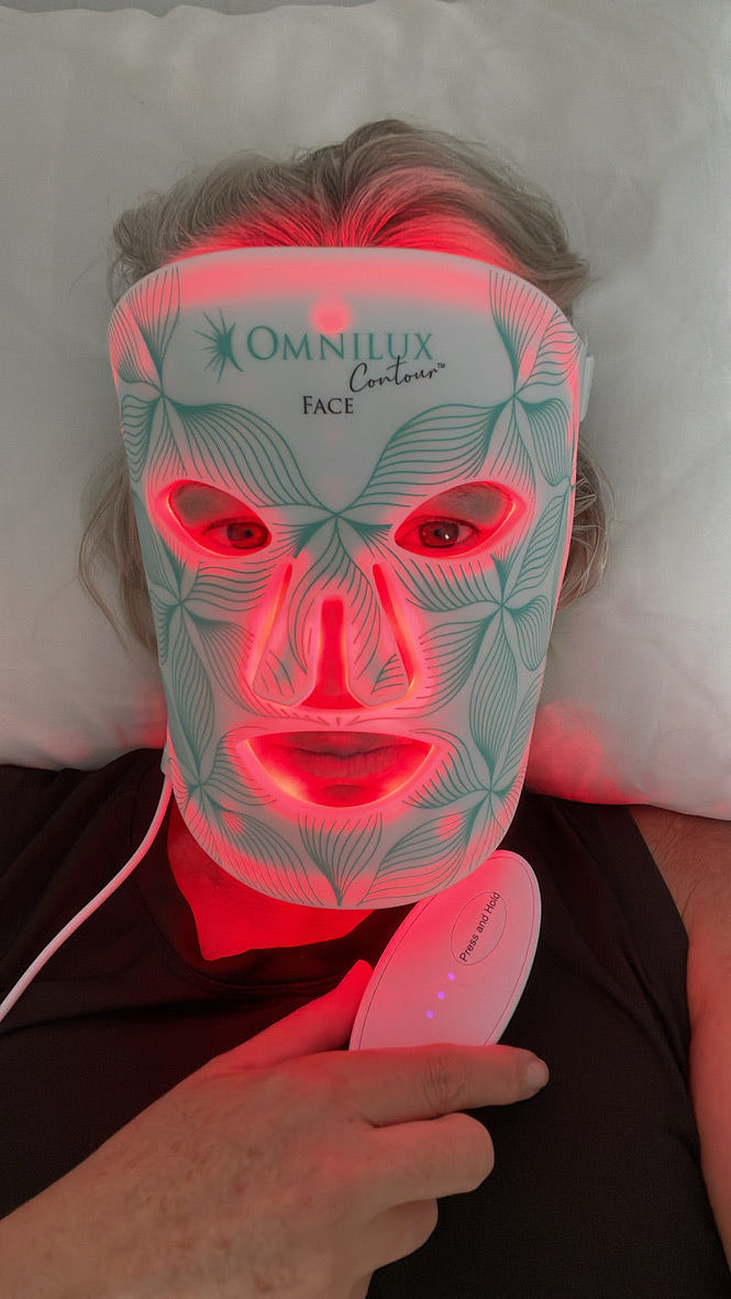 An over 40 year old woman lying down wearing an Omnilux Face LED light therapy mask that glows red.